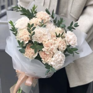 Bouquet of peach carnations with green greenery, wrapped in white translucent paper and tied with a peach ribbon.