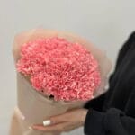 Pink carnation bouquet wrapped in light paper being held by a person in a black sleeve.