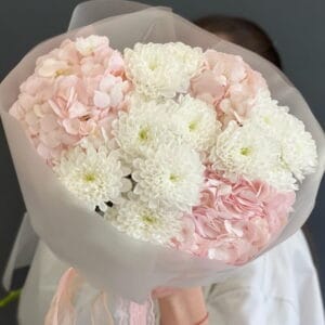 Bouquet of white chrysanthemums and pale pink hydrangeas wrapped in white paper, held by a person.