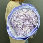 Person wearing a beige sweater holds a large lavender-wrapped bouquet of pale purple hydrangeas.
