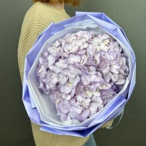 Person wearing a beige sweater holds a large lavender-wrapped bouquet of pale purple hydrangeas.